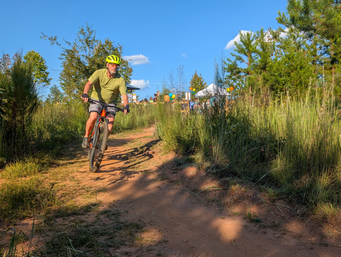 A person wearing a bright yellow helmet rides a mountain bike down a dirt trail surrounded by tall grass and trees. In the background, a gathering with colorful balloons and a tent is visible under a blue sky with a few fluffy clouds.