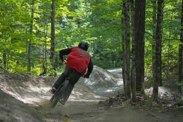 A mountain biker in a red vest navigates a dirt trail surrounded by lush green trees, leaning into a curve as dust kicks up from the bike's tires. The trail features jumps and is set in a vibrant, wooded area. Wilderness Tours Bike Park mountain bike trail.