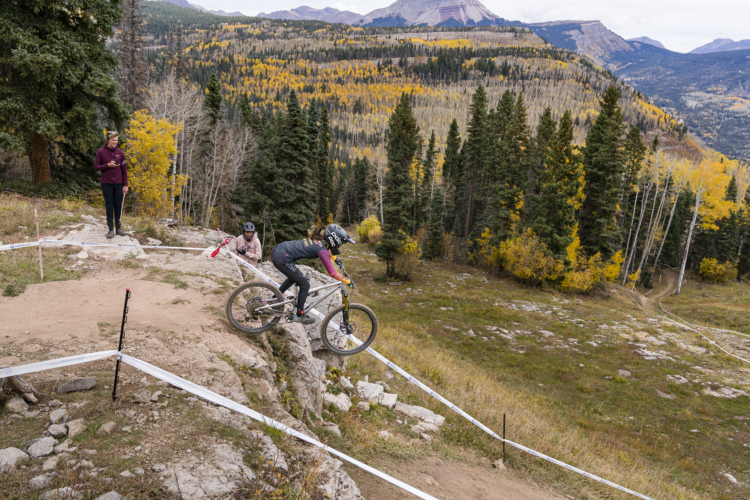 A mountain biker descends a rocky trail surrounded by autumn foliage, while two spectators watch from the side. The scene captures a vibrant landscape with trees displaying yellow leaves, steep mountains in the background, and race markers outlining the trail. The atmosphere conveys an adventurous, outdoor activity in a picturesque setting.