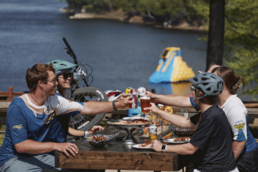 A group of cyclists enjoys a celebratory meal beside a lake, raising drinks in a toast. The table features a variety of food, including pizza and wings, and the scene is set in a sunny outdoor environment with bicycles nearby. Wilderness Tours Bike Park mountain bike trail.