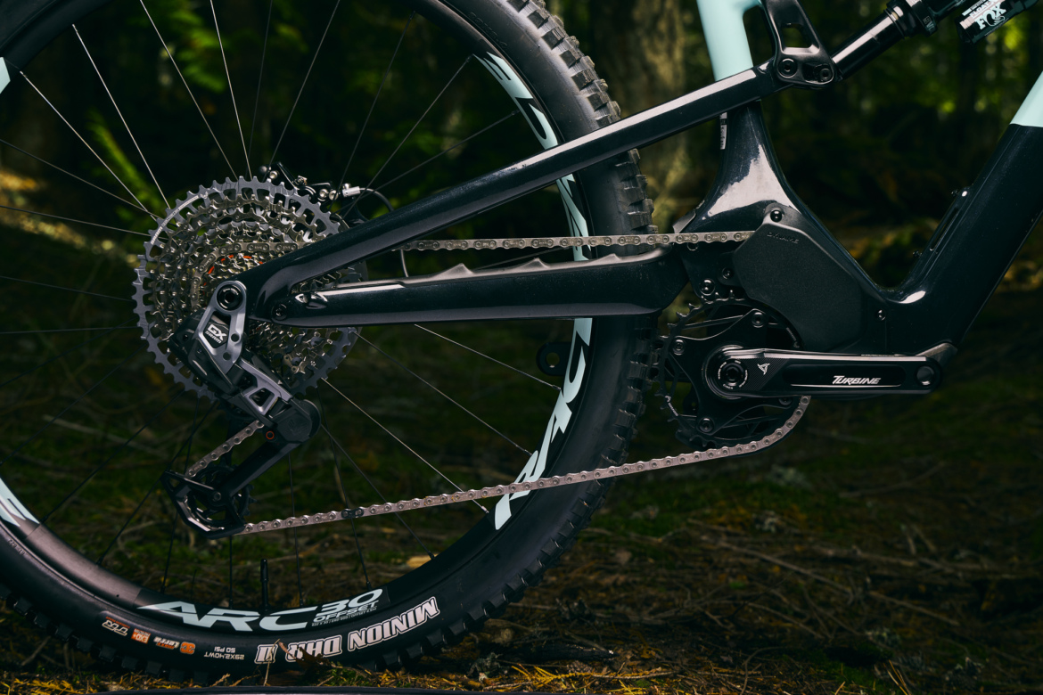 Close-up view of a mountain bike's drivetrain, featuring a black gear system, derailleur, and chain, with a backdrop of forested terrain and natural lighting. The tire visible is marked with "ARC 30" and exhibits rugged treads suitable for off-road biking.