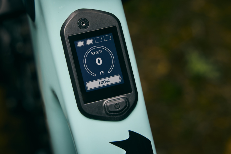 Close-up of a digital display mounted on a bicycle frame, showing a speedometer reading 0 km/h and a battery level indicator at 100%. The display features a sleek design with a circular speed gauge and icons in a modern aesthetic, set against a light blue frame background.