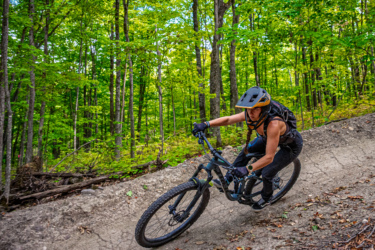 A person riding a mountain bike on a dirt trail surrounded by lush green trees and foliage. The cyclist is leaning into a turn, wearing a helmet and a backpack, showcasing an active outdoor lifestyle. Wilderness Tours Bike Park mountain bike trail.