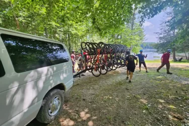A group of individuals near a trailer loaded with bicycles, set against a backdrop of trees and a lake. A white van is parked nearby, and the scene captures the excitement of preparing for a biking adventure. Sunlight filters through the leaves, creating a vibrant outdoor atmosphere. Wilderness Tours Bike Park mountain bike trail.