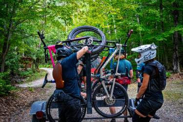 A group of mountain bikers preparing for a ride, with several bicycles loaded on a trailer. The scene is set in a lush, green forest area with a dirt path. One rider is adjusting a bike on the trailer while others are engaged in conversation, wearing helmets and biking gear. Wilderness Tours Bike Park mountain bike trail.