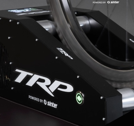 A close-up image of a stationary bike roller with a wheel in contact with the rollers. The device features the logo "TRP" prominently displayed on the front and includes a "powered by Sinter" label. The setup allows for indoor cycling training or testing.