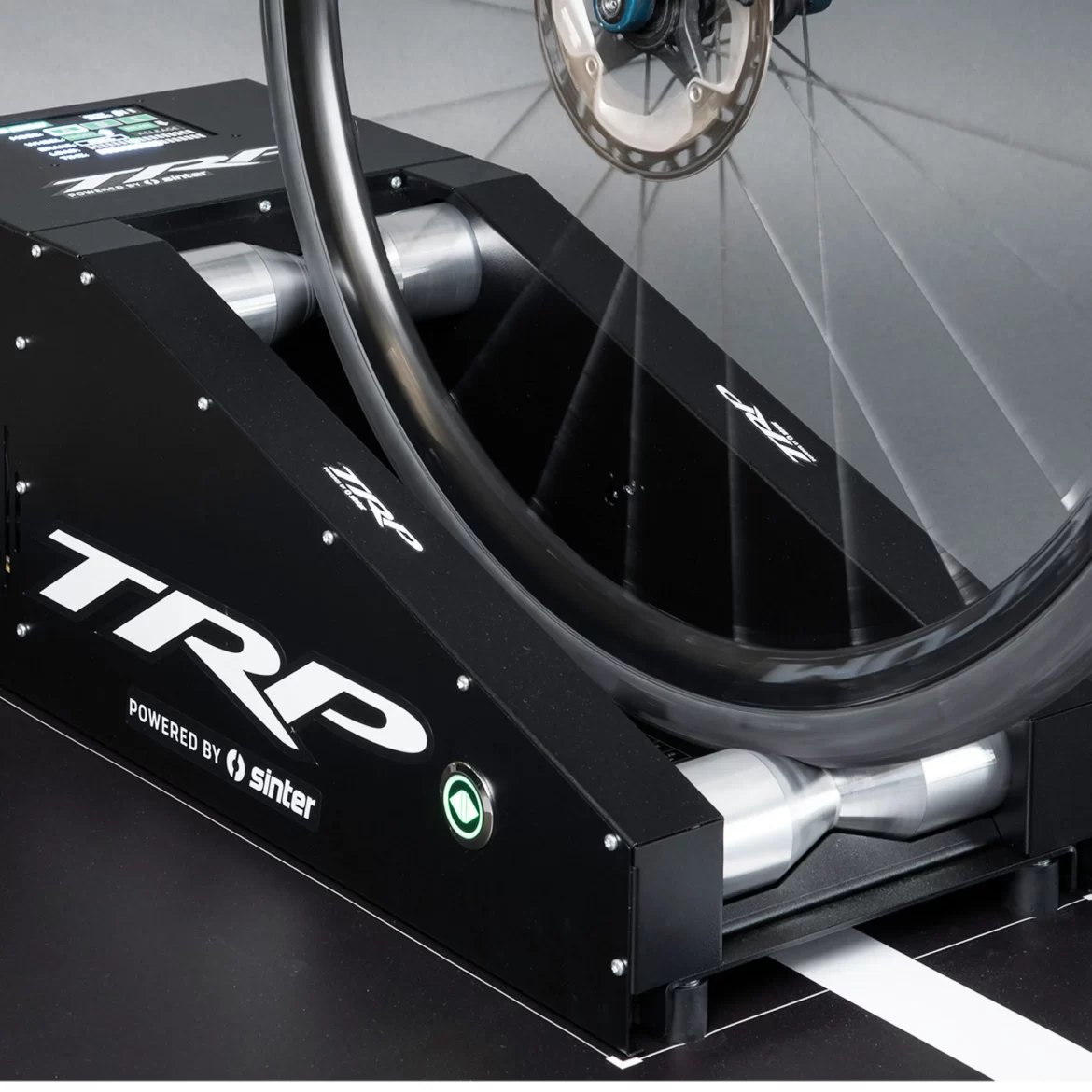 A close-up view of a bicycle on a black TRP trainer, featuring a digital display with various metrics. The bicycle wheel is in motion, and the trainer has metal rollers designed to assist in indoor cycling. The background is subtle and focuses on the equipment.