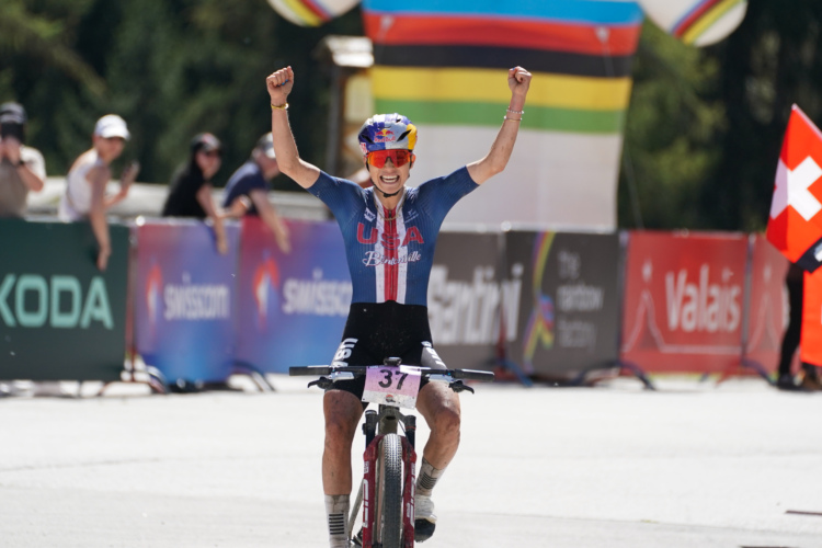 A cyclist celebrating triumphantly with both arms raised in the air, wearing a blue and red sports uniform marked "USA," crossed the finish line of a race. The background features a colorful finish line arch, with spectators visible in the distance, some holding flags and banners. The athlete appears joyful and victorious.