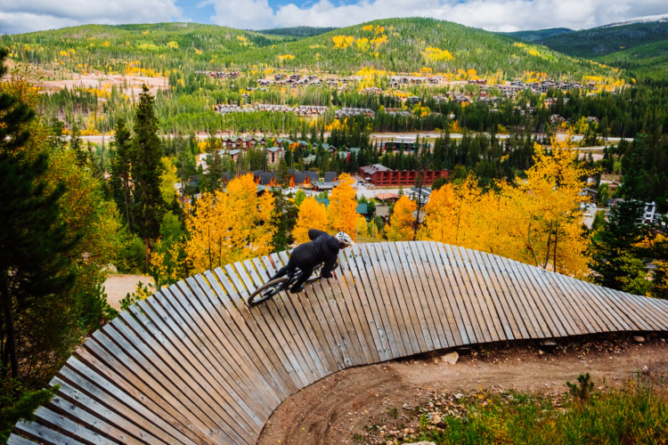 A mountain biker riding on a curved wooden trail with colorful autumn foliage in the background, showcasing vibrant yellow and green trees and a scenic view of a mountain village below.