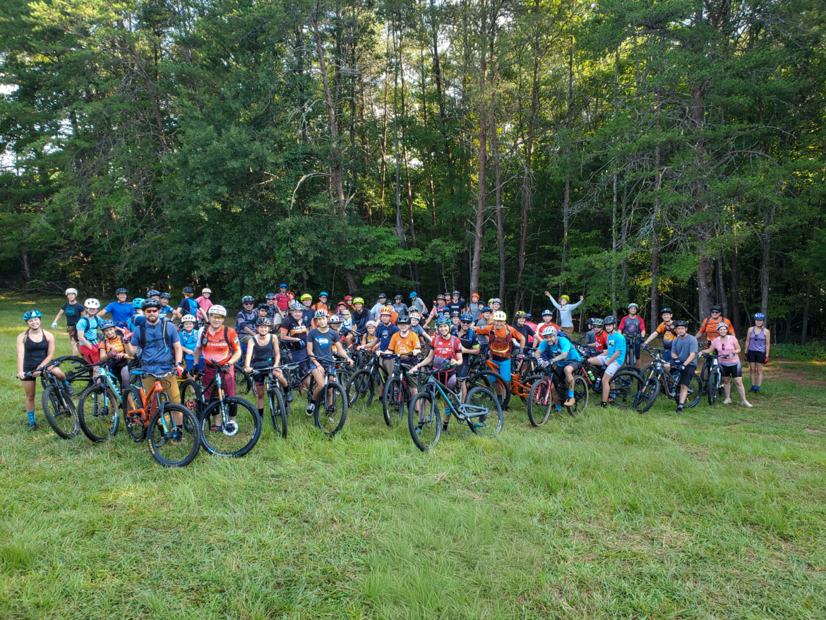 A large group of individuals posing on mountain bikes in a grassy area surrounded by trees. Participants are wearing helmets and varied cycling attire, with some people smiling and waving at the camera. The scene captures a sense of community and enthusiasm for biking outdoors.