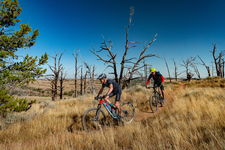 Two mountain bikers ride along a dirt trail surrounded by tall grasses and sparse, dead trees against a clear blue sky. The landscape features rolling hills in the background, suggesting a scenic outdoor environment ideal for cycling. One biker is dressed in a black jersey and shorts, while the other wears a bright red and black outfit with a yellow helmet.