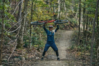 A mountain biker wearing a helmet and protective gear stands on a dirt trail in a forest, raising a red mountain bike above his head in a celebratory pose. Surrounding him are trees with green leaves, creating a vibrant, natural backdrop. Wilderness Tours Bike Park mountain bike trail.