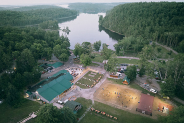 Aerial view of a lakeside retreat center surrounded by lush greenery. The image shows a cluster of buildings with green roofs, an outdoor gathering area, and a sports field illuminated for evening activities. A serene lake and wooded hills are visible in the background. Wilderness Tours Bike Park mountain bike trail.