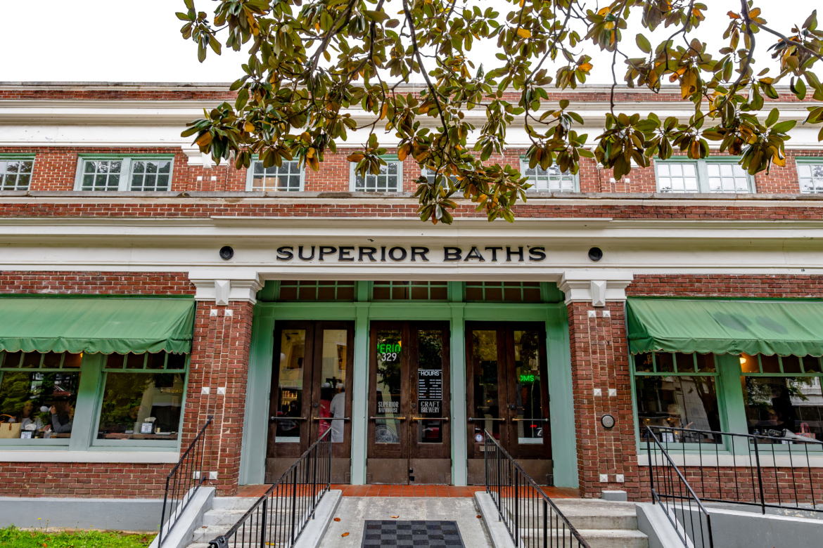 Image of the entrance to Superior Baths, a brick building with green awnings and large glass doors. The business name "SUPERIOR BATHS" is displayed prominently above the entrance. The scene includes steps leading up to the door and a nearby tree with green foliage.