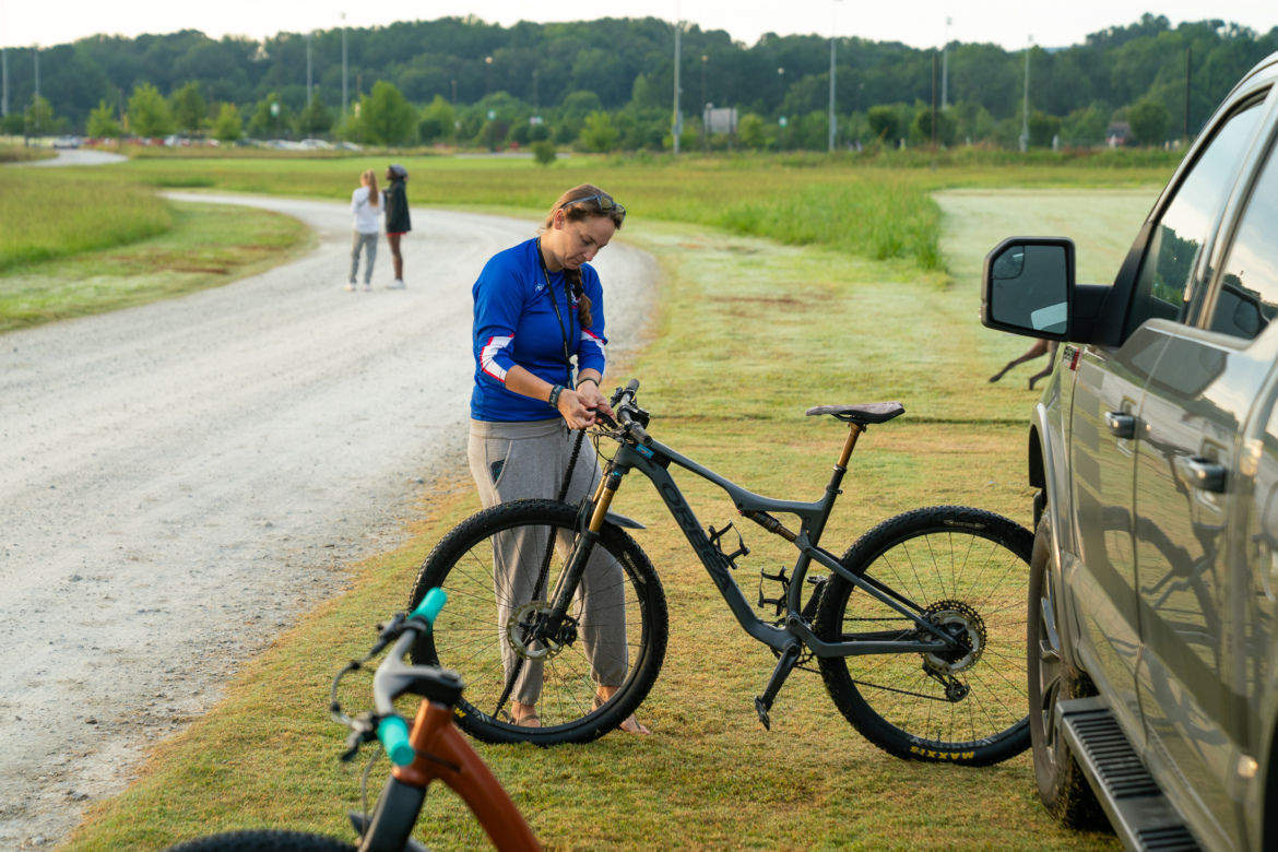 A woman stands beside a parked truck, adjusting her mountain bike on a grassy area near a winding dirt path. In the background, two people are walking along the path, surrounded by green trees. The early morning light creates a serene atmosphere.