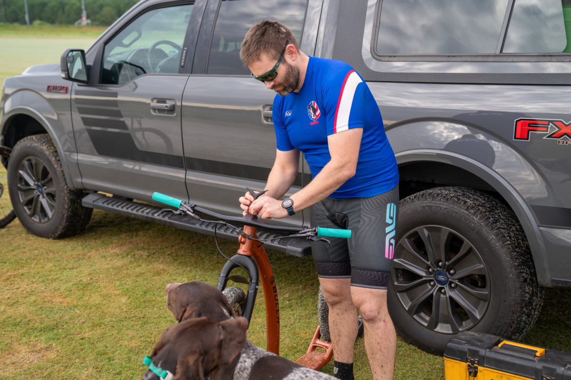 A person in a blue athletic shirt and cycling shorts is working on a scooter while standing next to a gray pickup truck. Two dogs are nearby, looking up at the person. The setting is outdoors on a grassy area, and the scene suggests an active lifestyle.