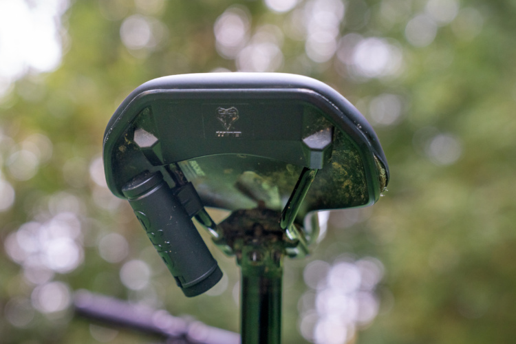 Close-up view of a metal detector's search coil, with a focus on its underside. The coil is black, showing some signs of wear and dirt, and features attachment points for a shaft. A blurred, green and natural background suggests an outdoor setting.