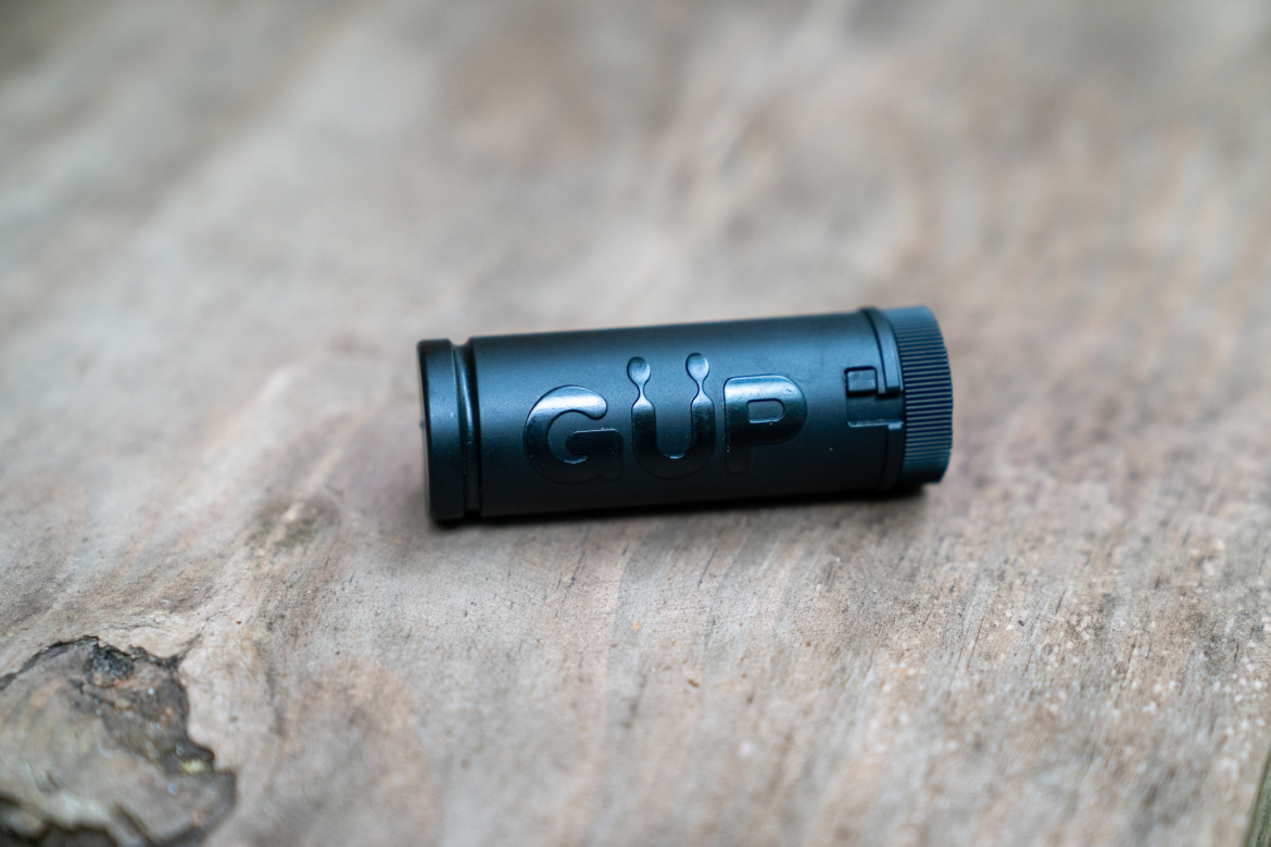 A close-up image of a black cylindrical device labeled "GUP," placed on a wooden surface. The device features a textured end and a sleek design, with the logo prominently displayed.