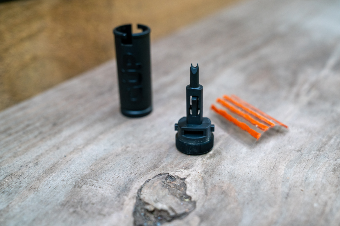 A close-up image of a black tool component resembling a valve, accompanied by a cylindrical casing and three thin orange strips, resting on a rustic wooden surface.