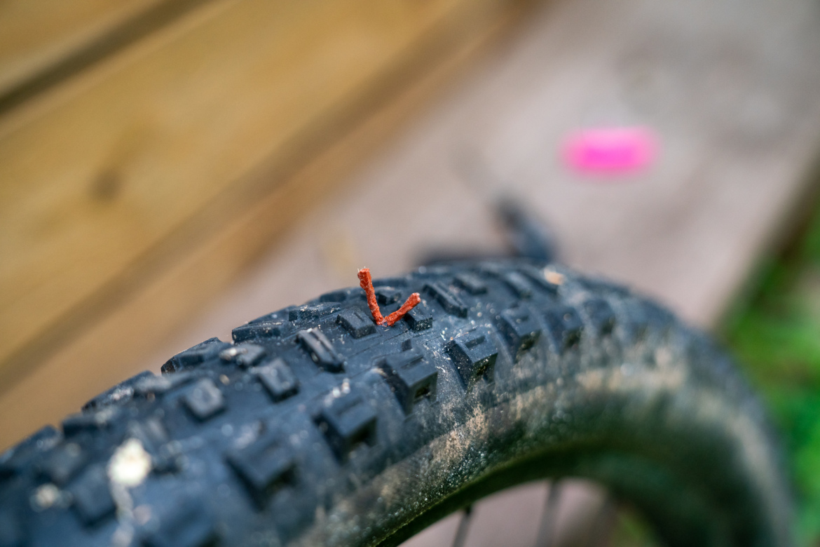 A close-up image of a bicycle tire showing a small piece of reddish-brown rusted metal embedded in the tread. The background is slightly blurred, revealing wooden planks and a hint of pink object.