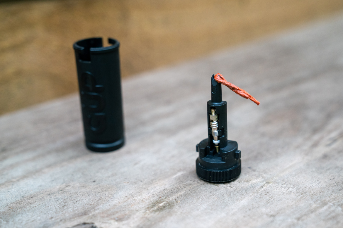 A close-up image of a black bicycle valve tool with a bright orange string attached, placed on a wooden surface. Next to it is a cylindrical black container, partially visible. The background features a blurred, neutral-colored surface.