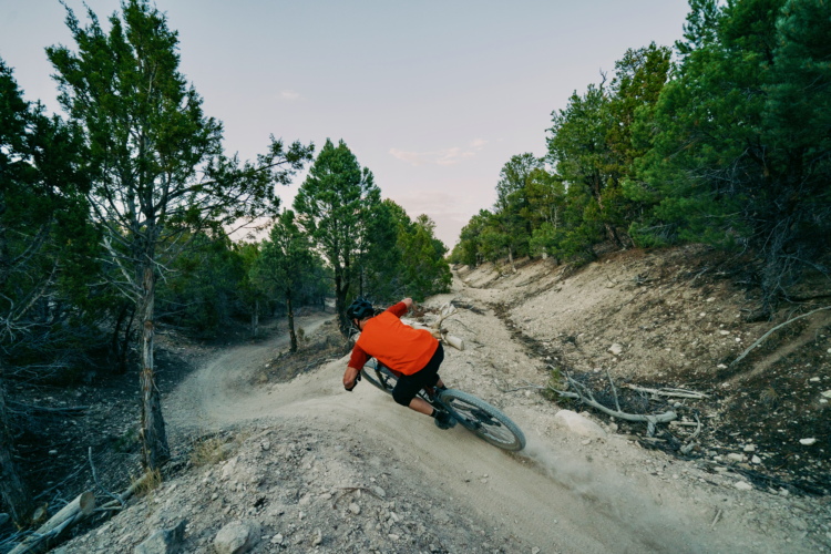 A mountain biker in an orange shirt leans into a turn on a dirt trail surrounded by trees, kicking up dust as he navigates a winding path through a mountainous landscape.