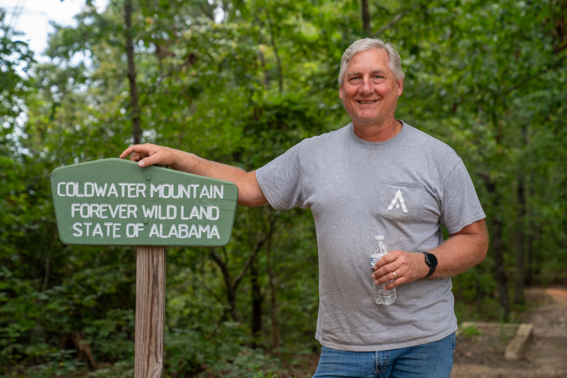 Marcus Tillman, Recreation Trail Director for the city of Anniston, Alabama, stands beside a sign that reads "Coldwater Mountain Forever Wild Land State of Alabama." He is smiling and holding a water bottle, with a background of lush green trees and a hiking path.