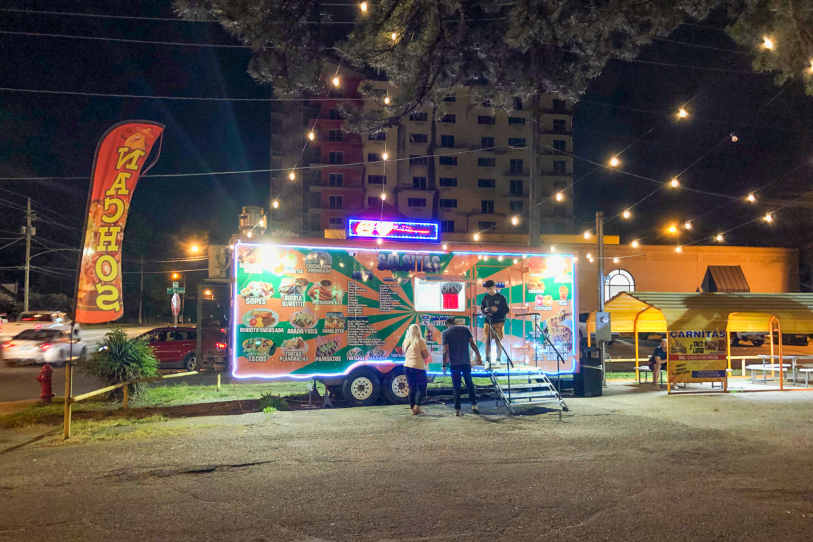 Brightly lit food truck at night with a colorful menu displayed, where two customers are ordering food. A large banner with the word "Sopas" is visible, along with several food options like tacos and burritos. Surrounding the truck, there are string lights and nearby buildings in the background.