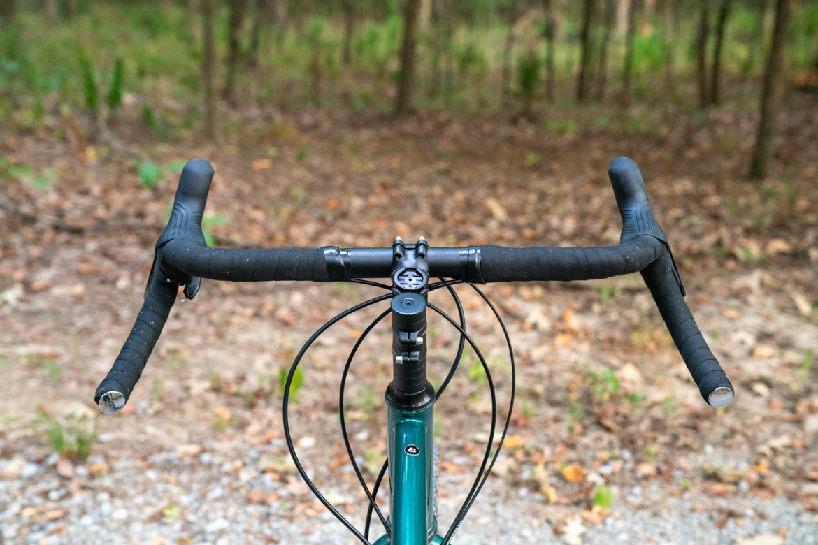 Close-up view of a bicycle handlebar set against a blurred natural background, with the bike's stem and cables visible. The handlebars are wrapped in black tape, and the scene conveys a sense of outdoor adventure.