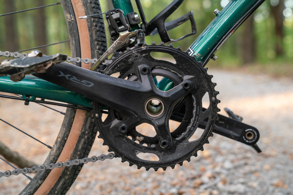 Close-up of a bicycle's crankset and chain, highlighting the black chainrings and pedals, with a backdrop of a gravel path and blurred greenery.