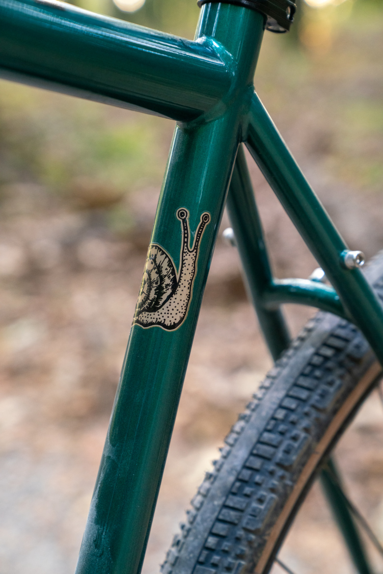 Close-up of a green bicycle frame featuring a decorative snail illustration on the downtube, with a blurred natural background.