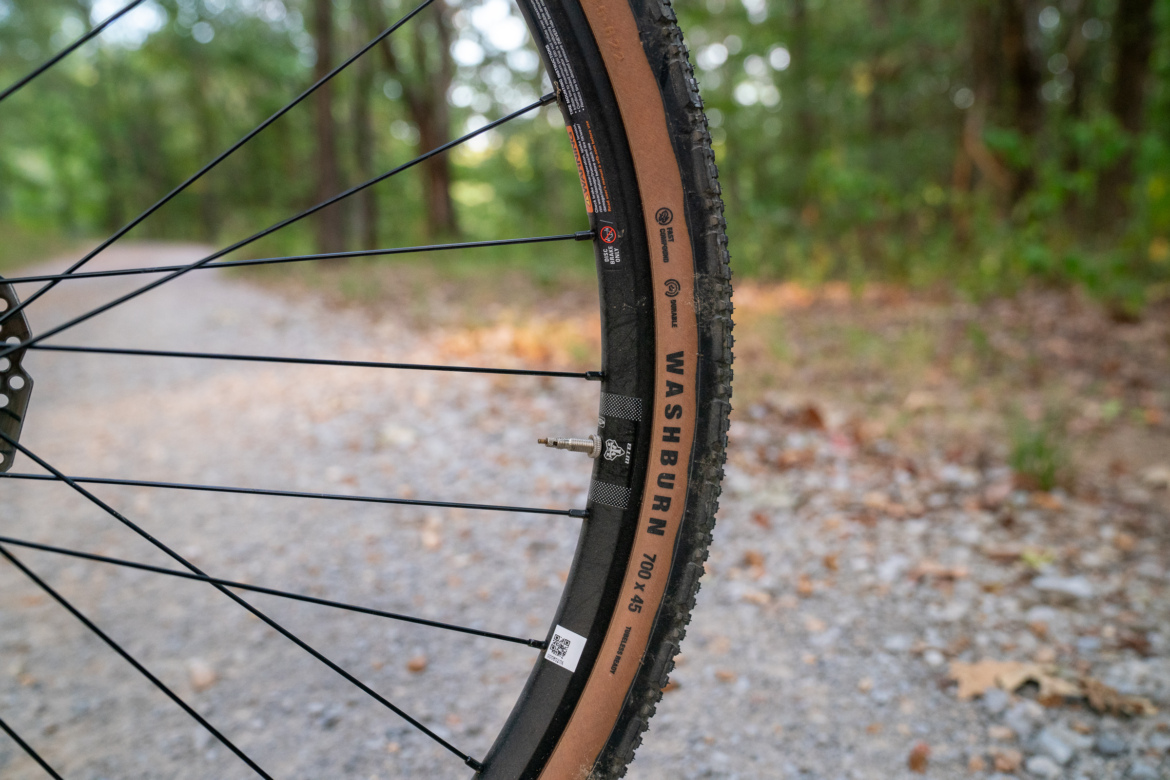 Close-up of a bicycle tire on a gravel path, featuring a brown and black design with the word "WASHBURN" visible on the sidewall. The background shows blurred greenery, indicating a natural outdoor setting.