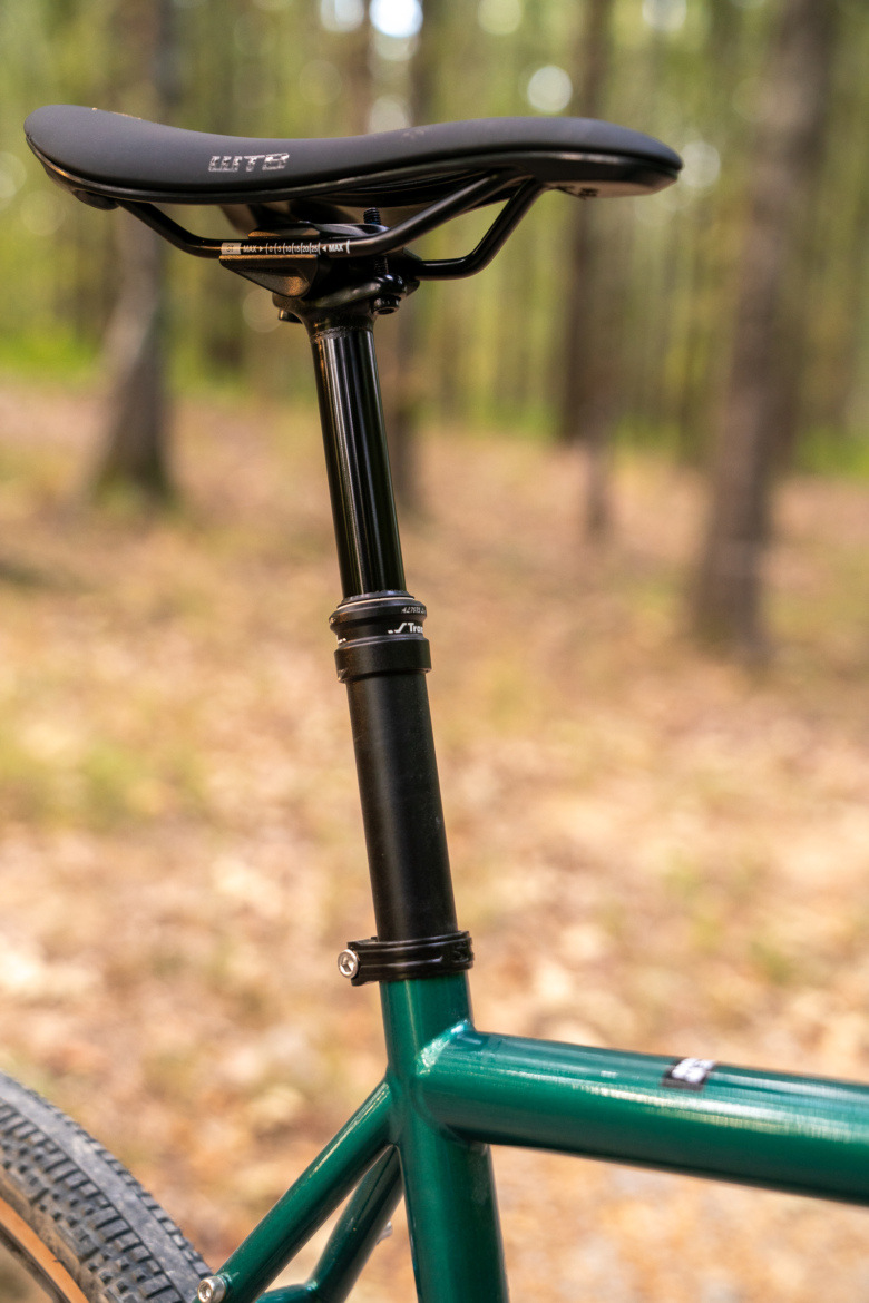 Close-up image of a bicycle saddle and seat post, featuring a black WTB saddle mounted on a sleek black seat post, attached to a green bike frame. The background shows a blurred natural setting with trees and foliage.