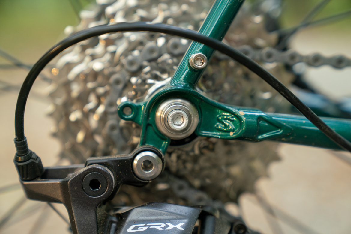 Close-up image of a bicycle's rear derailleur and cassette, showing a green frame component, silver gears, and exposed chain. The focus is on the mechanical details, highlighting the components involved in gear shifting.