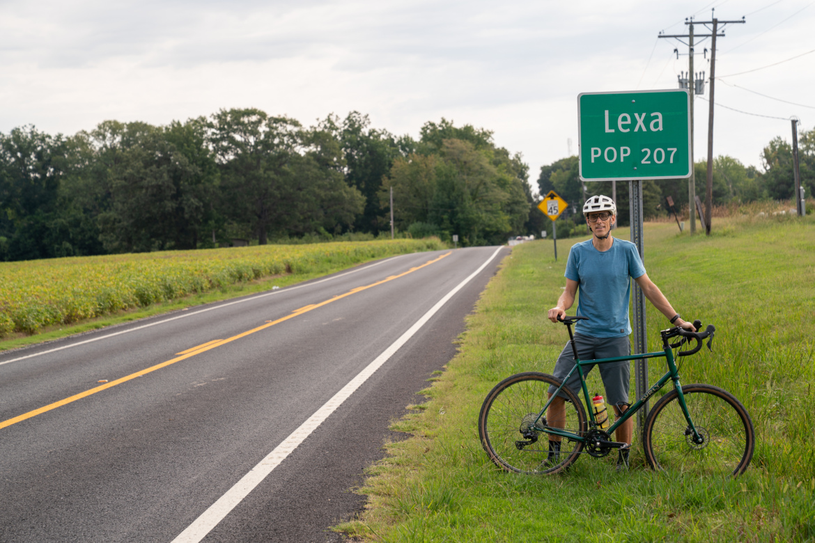 A person in a blue t-shirt and a helmet stands next to their bicycle beside a highway sign that reads "Lexa POP 207." The scene features a rural road, lush green fields, and a cloudy sky in the background.