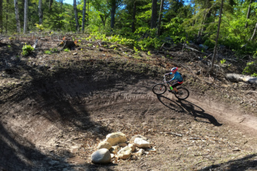 A child riding a mountain bike on a dirt trail surrounded by trees, with a circular dirt feature and a few rocks in the foreground. Sunlight is filtering through the leaves, creating a vibrant and active outdoor scene. Palmer Woods Forest Reserve mountain bike trail.