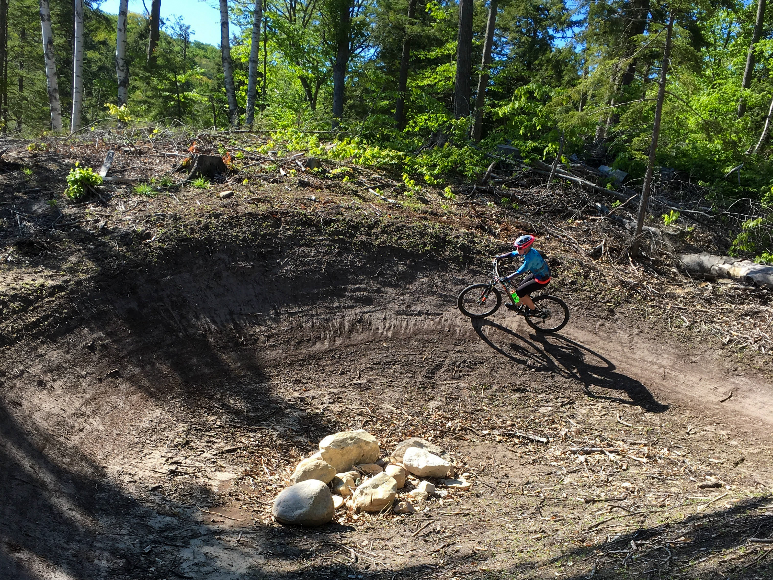 A child riding a mountain bike on a dirt trail surrounded by trees, with a circular dirt feature and a few rocks in the foreground. Sunlight is filtering through the leaves, creating a vibrant and active outdoor scene. Palmer Woods Forest Reserve mountain bike trail.