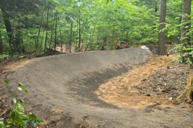 A curved dirt track in a forested area, surrounded by lush green trees and underbrush. The surface appears freshly groomed, with a smooth, compact texture. In the background, a small piece of heavy machinery is partially visible, indicating ongoing construction or maintenance work. Palmer Woods Forest Reserve mountain bike trail.