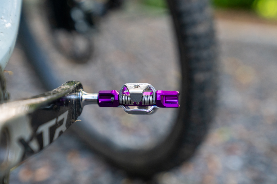 Close-up view of a bicycle pedal featuring a purple clip mechanism, mounted on a crank arm. The background shows the tread of a bike tire and a gravel surface, suggesting an outdoor setting.