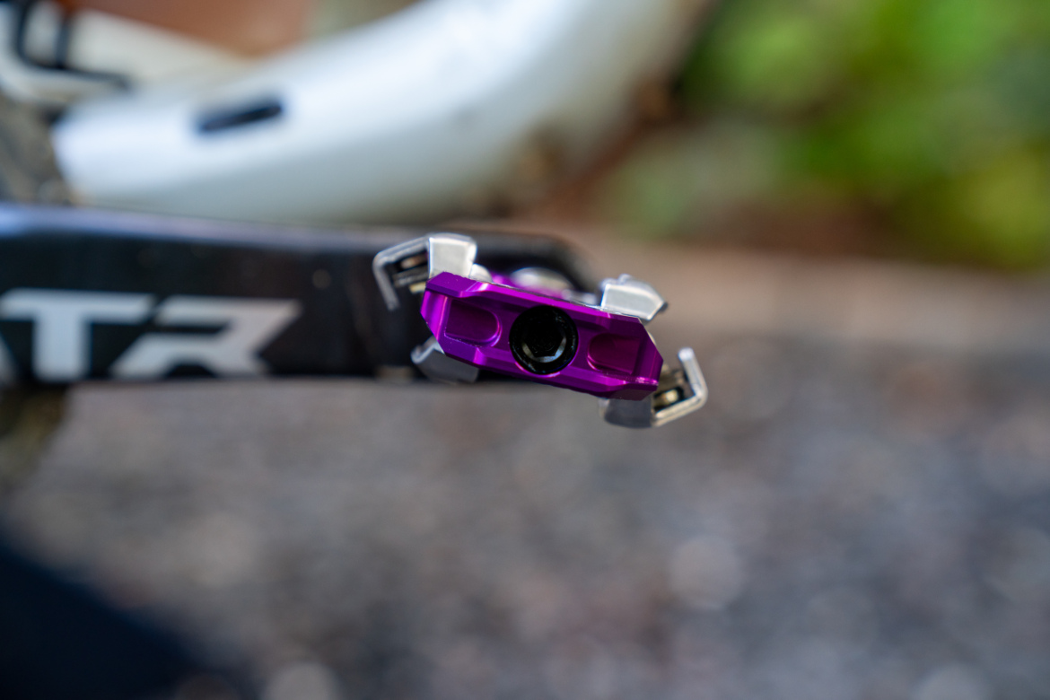 Close-up image of a purple bicycle pedal attached to a bike crankarm, showing its intricate design and metallic finish. The background features blurred greenery, indicating an outdoor setting.