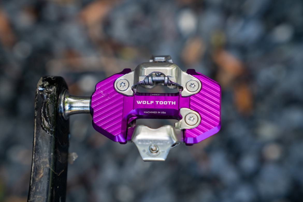 An overhead view of a purple mountain bike pedal featuring a silver metallic mechanism. The pedal has a textured surface and is marked with the brand name "WOLF TOOTH" and "MACHINED IN USA." The background consists of blurred gravel and fallen leaves, creating an outdoor setting.