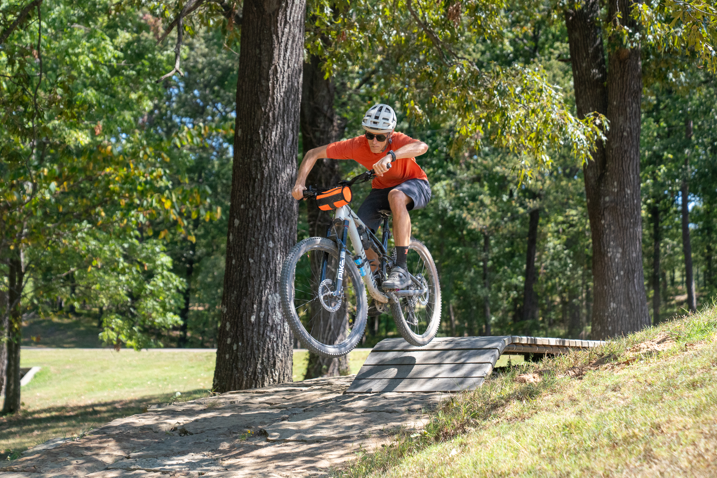 A young person wearing an orange shirt, shorts, and a helmet is jumping off a wooden ramp on a mountain bike in a forested park. Trees with green leaves surround the area, and the rider is in mid-air, showing an athletic pose. The bike features a vibrant orange bag on the front. Sunlight filters through the trees, illuminating the scene.