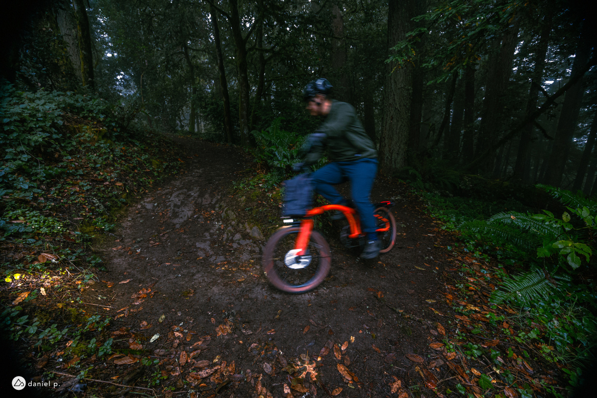 A person riding an orange bicycle on a muddy trail surrounded by dense greenery and trees in a misty forest. The cyclist is in motion, creating a sense of speed as they navigate the winding path covered with fallen leaves.