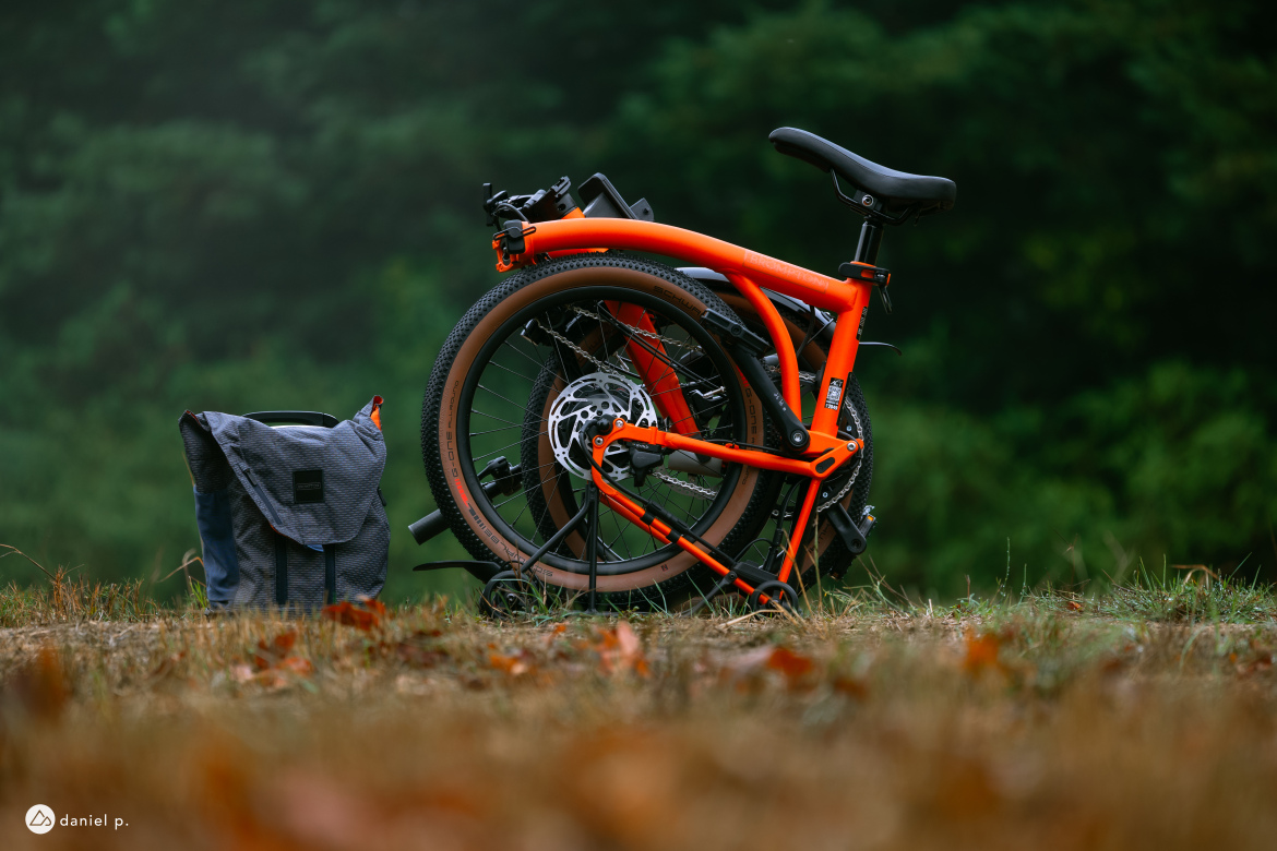 A folded orange bicycle is positioned next to a gray messenger bag on grassy terrain, with a blurred background of green trees. The bicycle is compact, with the seat up and the wheels secured, indicating it's designed for portability. The scene is set in a natural outdoor environment, suggesting an adventure or travel theme.