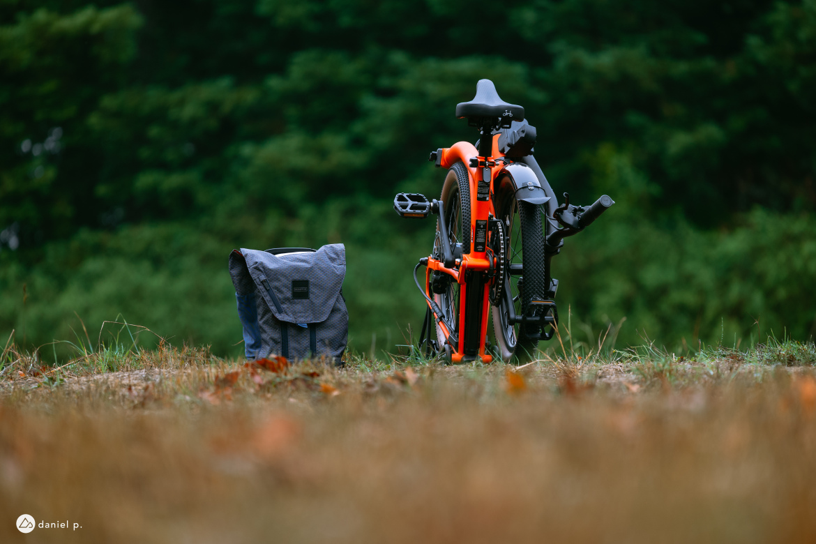 A folded bicycle in bright orange and black parked on a grassy area, next to a gray backpack. The background features blurred greenery, suggesting an outdoor setting.