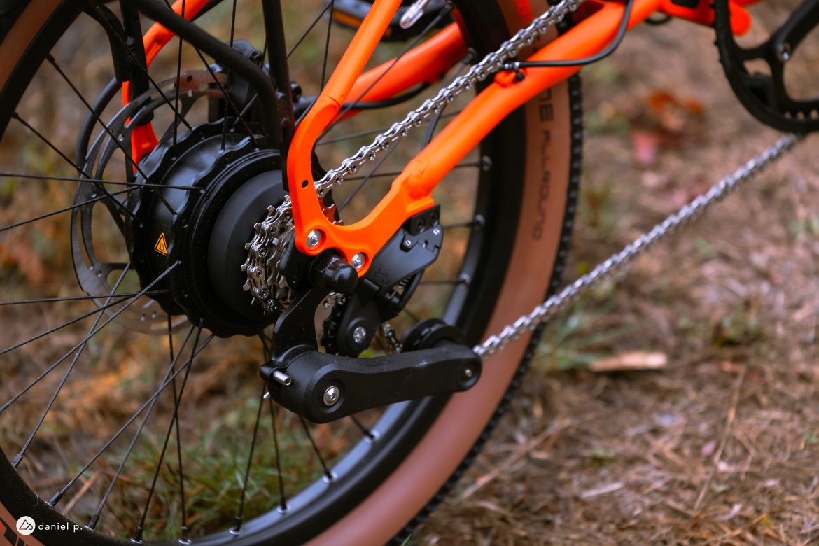 Close-up view of the rear wheel and drivetrain of a bicycle, featuring a vibrant orange frame, a black hub motor, and a chain connected to the gear system. The background shows earthy tones with some grass and leaves.