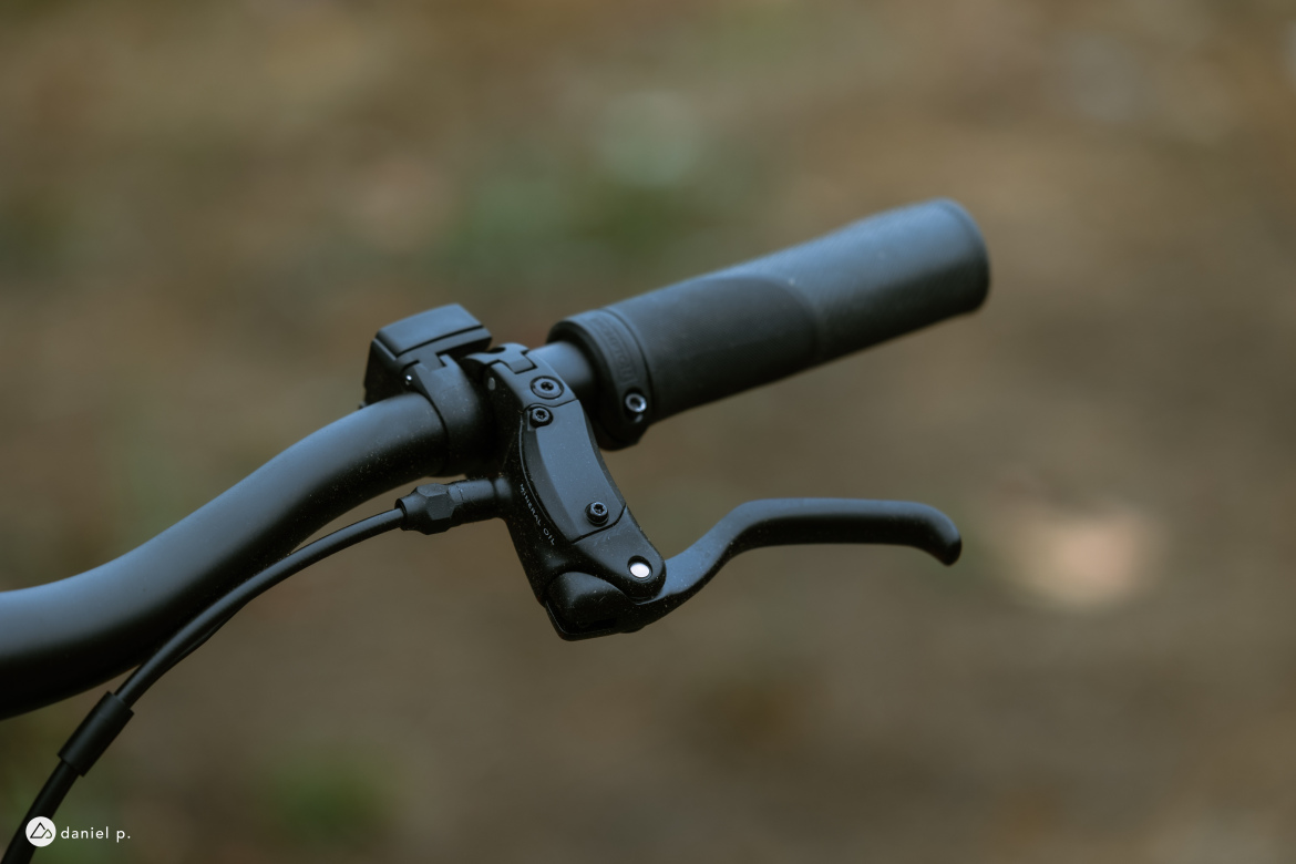 Close-up image of a mountain bike handlebar featuring a black brake lever, a textured black grip, and visible cables. The background is blurred with earthy tones, suggesting an outdoor setting.