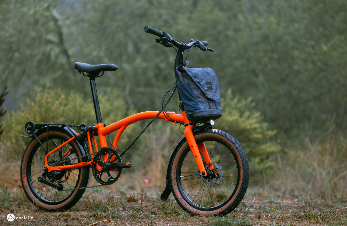 A vibrant orange folding bicycle parked in a natural setting, featuring a blue bag attached to the rear. The background includes soft greenery and a blurred natural landscape.