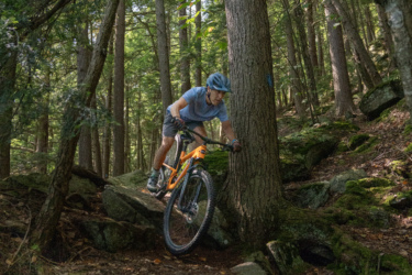A mountain biker navigates rocky terrain in a dense forest, leaning forward as they approach a tree, with sunlight filtering through the leaves above. The biker wears a helmet and casual athletic clothing while riding a bright orange mountain bike. Leicester Hollow and Chandler Ridge Loop mountain bike trail.