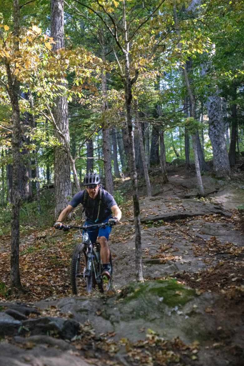 A mountain biker navigates a rocky trail in a forested area, surrounded by trees with autumn foliage. The rider, wearing a helmet and cycling gear, appears focused as they maneuver over the uneven terrain. Sunlight filters through the leaves, creating a lively and dynamic atmosphere.